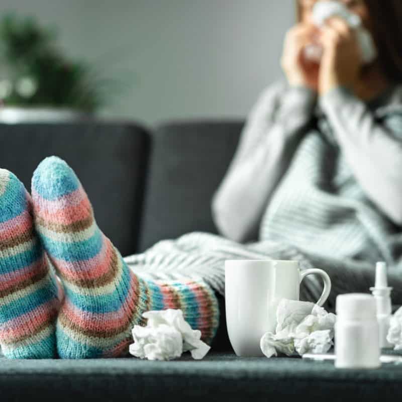 Sick woman with flu or covid-19 with fever and cough sitting on couch at home. Ill person blowing nose and sneezing with tissue and handkerchief. Woolen socks and medicine. Infection in winter. Resting on sofa.