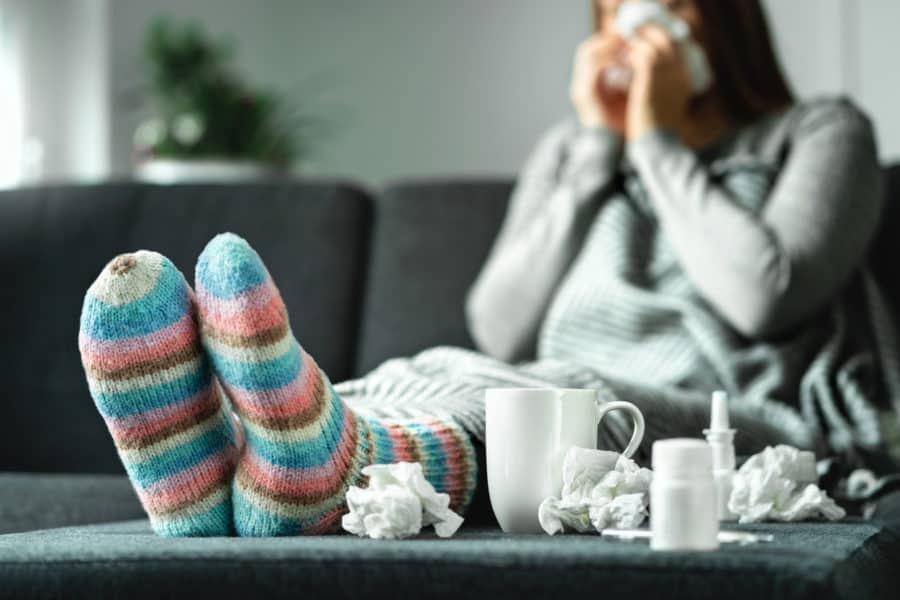 Sick woman with flu or covid-19 with fever and cough sitting on couch at home. Ill person blowing nose and sneezing with tissue and handkerchief. Woolen socks and medicine. Infection in winter. Resting on sofa.