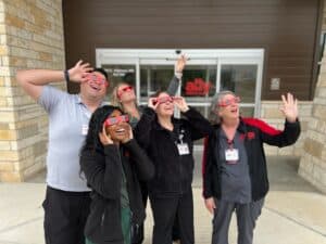 The Ally Medical staff, adorned with eclipse glasses, stand together outside the ER building, embodying a sense of unity and excitement. 