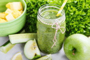 A vibrant green apple smoothie in a clear glass, garnished with a slice of green apple on the rim, surrounded by fresh apples and spinach leaves, embodying the spirit of a healthy, family-friendly St. Patrick's Day.