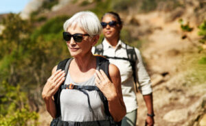 Two elderly women hiking together on a scenic trail, smiling and enjoying the outdoors. They are dressed in comfortable hiking gear, surrounded by lush greenery, symbolizing active and healthy aging.